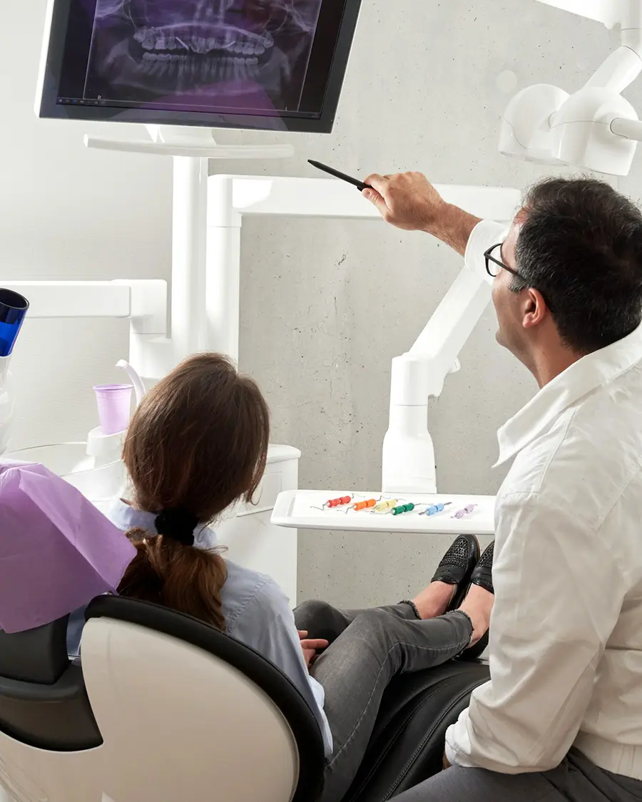 A dentist at a dental practice that found the most trusted group health insurance agency in Texas points at a dental X-ray on a monitor, explaining it to a patient seated in the chair. Dental tools and a cup are visible nearby.